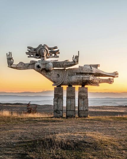 Monument "Peace" by Georgian sculptor Nugzar Manjaparashvili, created in the 1970s, stands against a stunning sunset in Nukriani, Georgia. The brutalist-style sculpture depicts a stylized female figure in a floating posture, supported by four massive concrete pillars. With arms outstretched and an expressive face framed by an intricate headdress, the figure appears both serene and commanding. The surrounding landscape of rolling hills and distant mountains enhances the monument’s dramatic presence. Captured in 2024 by photographer Stefano Perego.