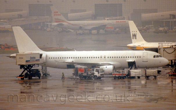 Side view of a white, twin engined jet airliner parked facing to the right.
There is a hatch open in the lower forward fuselage, with a white luggage belt raised up to load bags into the plane.
Baggage carts are parked in front of the plane's nose.
A door is open in the rear fuselage, with the body of a white truck with blue "Dobb's" titles on the side raised up to allow access through this door.
A fuel truck is parked by the side of  the plane's right engine.
A white airbridge is attached to the forward door on the far side of the plane.
In the background, the tail of a white and yellow jet airliner is parked facing to the right on the right of the frame.
In the background, a white, twin engined jet airliner with a red and gold stripe running along the body and red "Air 2000" titles on the upper forward fuselage and tail, is parked in front of a grey terminal building that stretches across the frame, slowly being buried in mist.