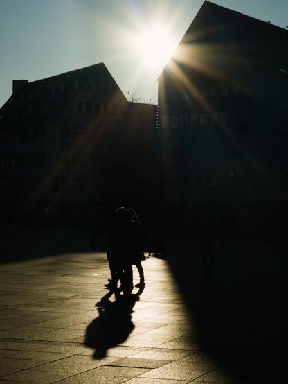 A silhouette of two people, walking closely together on a patch of light. The scenery is the minster place of Ulm. There are two houses in the background, also mostly silhouetted by the descending sun standing just above the roofs.The rest of the images fades into black.