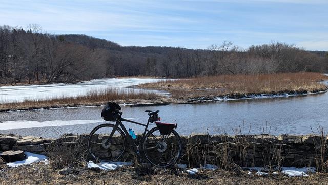 Dark brown Diverge bicycle with Bar Mitts leaning against a short stone wall in front of Mayowood Lake looking Southwest. The near part of the lake/river is open, the far part is still frozen. There are hills with bare trees further behind under mostly blue skies.