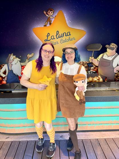 Two women posed in front of a La Luna themed midway game. One wearing a bright yellow dress and holding a star plushie and the other wearing brown overalls holding a Bambino plushie.