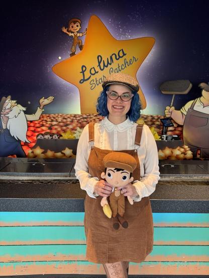 Woman wearing a pair of brown overalls and a hat holding a bambino plushie in front of a La Luna themed midway game.