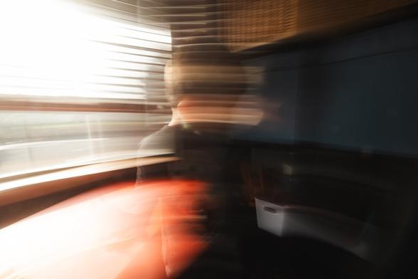 A more abstract photo utilizing a shutter drag, causing a lot of blur from the highlights to bleed through the image. A young child stands in center frame, a bright window behind them and a red table under the window. In front of them is a dark bookshelf. The window and tables brightness is bleeding over the image