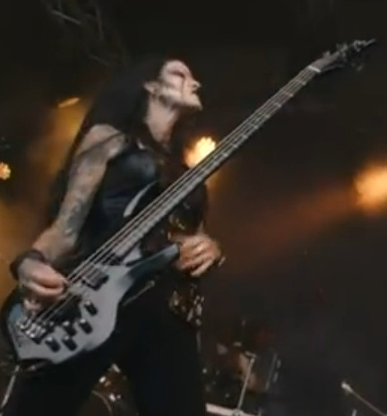 Young woman wearing so called corpse paint (face mostly white with black accents) and playing a bass guitar at a heavy metal concert, several tattoos are on her right arm
Due to the photographer's angle and the light effects, the overall atmosphere of the image is a bit surreal and mysterious