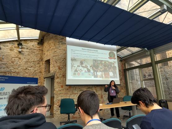 foto di una sala durante presentazione di slides proiettate, presentatrice sotto il telo che parla al microfono, persone sedute nelle prime file
