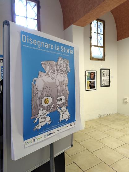foto del manifesto della mostra d'illustrazione chiamata "Disegnare la Storia" su uno stand all'interno di una sala, che si intravvede nello sfondo con finestre tra sezioni arcate e quadri appesi alle pareti sottostanti, ambienti usati anche per le conferenze del convegno CAST