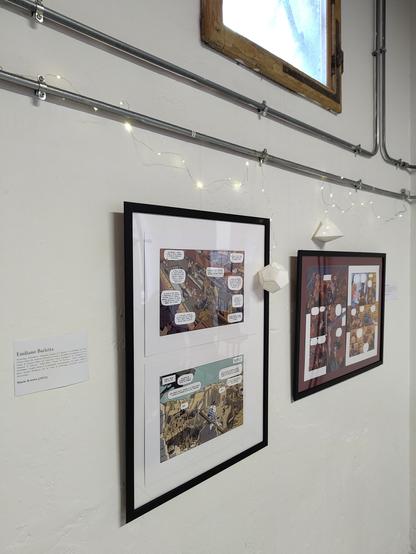 foto di due cornici espositive, che contengono le copie a stampa di tavole illustrate per vignette a tema storico-archeologico, appese su parete bianca sotto una finestra; cartellini esplicativi in forma di fogli di carta corredano i lati delle cornici e su quello più vicino si leggono in grassetto il nome dell'autore "Emiliano Barletta" e il nome dell'opera "Diario di scavo (2021)"
