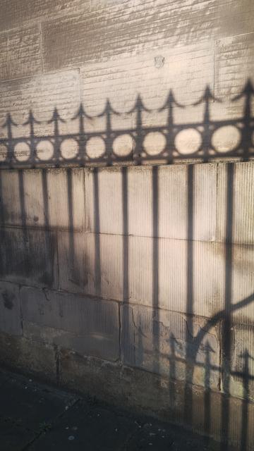 Sharp shadows of arrow headed railings cast onto a stone wall