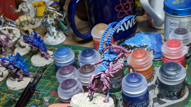 A Tyranid with big claws from the other side painted in blue and purple. In the background you can see many paint pots.