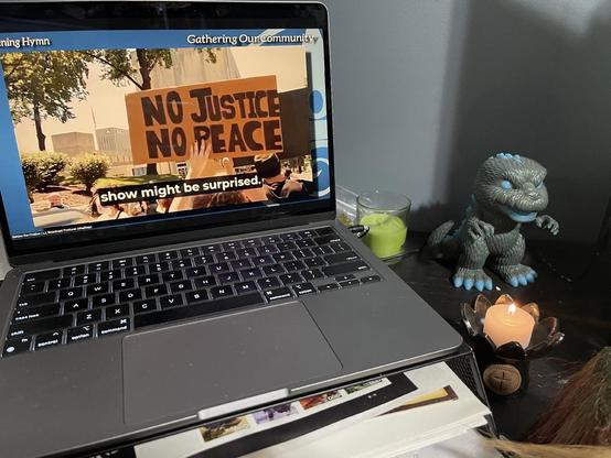 A laptop displays a protest scene with a sign that reads "NO JUSTICE NO PEACE," featuring subtitles. Nearby, a green candle and a decorative Godzilla figure sit on a desk, alongside a lit candle in a flower-shaped holder.