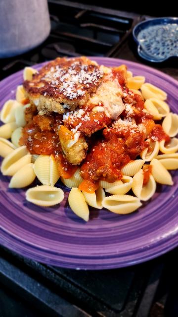 Purple dinner plate covered in pasta shells, red sauce, and a piece of breaded chicken coated with more sauce, cheese, and lastly a round mozzarella stick balanced on top, with a sprinkling of parmesan over everything.