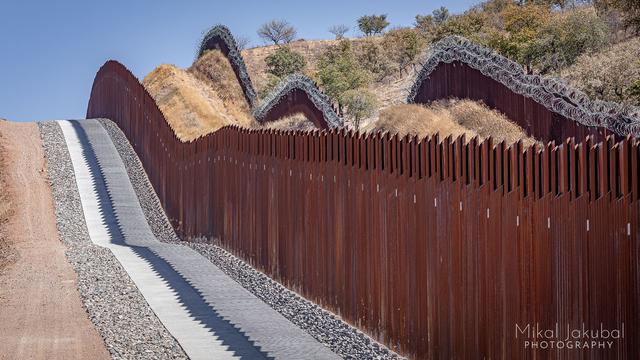 The cement border road, the new border fence and the old border fence next to that, all running in parallel up and over a few small hills before disappearing out of sight over a larger hill. The new fence is rusty steel. Then there is a wide, dry grassy area, then the old fence made of steel poles and covered with double layers of concertina wire.