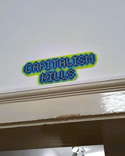 The words "CAPITALISM KILLS" made out of Hama beads. The beads are neon green and blue.