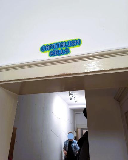 The words "CAPITALISM KILLS" made out of Hama beads, hung over a door frame. The beads are neon green and blue.