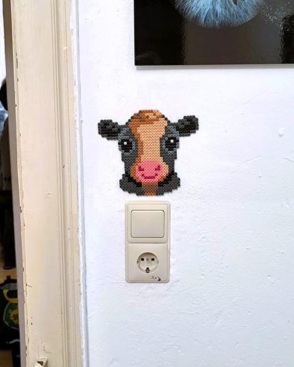A baby cow face made out of Hama beads. The cow is placed over a light switch on the wall.