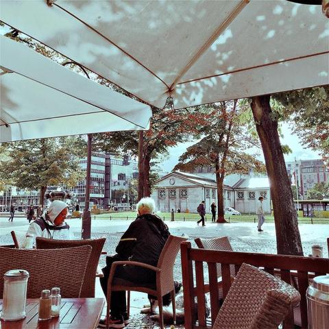 This picture shows a woman sitting at an outdoor table under a white umbrella next to a tree on a sunny day in the city. She is wearing a light-colored dress, sunglasses, and has long hair tied back. The background features buildings and trees. A woman in a black shirt and jeans walks by in the distance.