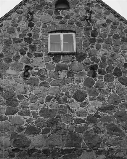 The image shows a stone wall of a building with a window near the top. The wall is constructed of irregularly shaped stones with visible mortar. There are some decorative elements on the wall, including a couple of horn-like features and some circular objects. The window has a brick frame and is divided into smaller panes.