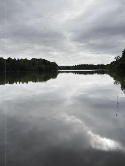 The image shows a calm lake with a reflection of the cloudy sky above. The lake is surrounded by dense, green trees. The overall atmosphere is serene and tranquil, with overcast skies indicating a possibly gloomy or calm day.