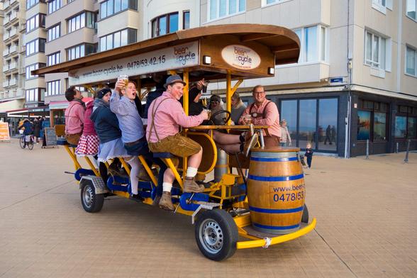 A group of people are riding a yellow beer cycle on a brick road. The beer cycle has a wooden roof with the words "beer cycle" on it. The people are sitting on seats and pedaling. Some of the people are holding beers. The person at the front is steering with a steering wheel. There is a wooden barrel at the front of the beer cycle. There are buildings in the background. There are also people walking in the background.