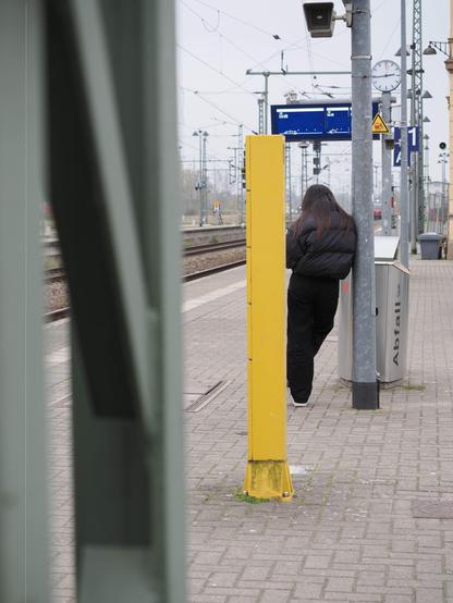 The image shows a train station platform. There is a person standing near a utility pole. The platform has various signs, a clock, and an overhead display board indicating train times. There are overhead electric lines and tracks visible in the background. A yellow pillar and a trash can labeled "Abfall" are also present.