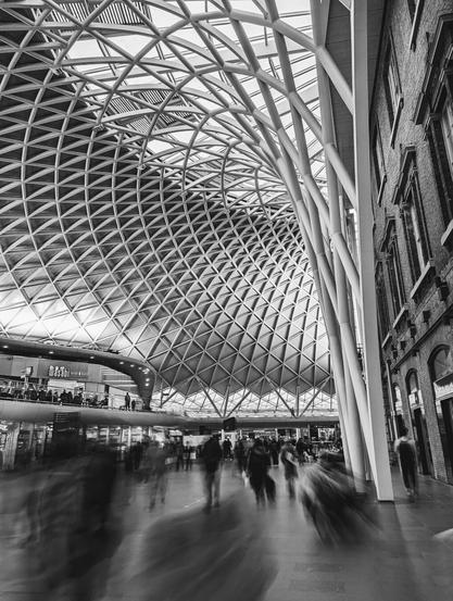 A black and white photograph of the interior of King's Cross Station in London. The dominant feature is the modern, curved, grid-like roof structure made of white beams, contrasting with the traditional brick building on the right. Below, blurred figures of people walk through the station, suggesting movement and activity.