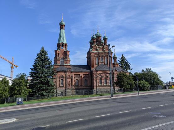 La cathédrale orthodoxe de Tampere, vue de côté. Elle est en brique rouge, toitures vertes sur les clochers et noire sinon, croix dorées sur les sommets. L'église est entourée d'un parc arboré. Au premier plan, une rue déserte. Au fond à gauche, une grue. Le ciel est bleu, très légèrement voilé par endroits.