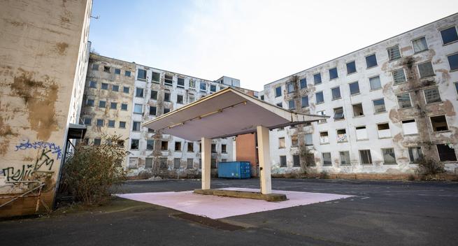 An abandoned petrol station canopy stands in the foreground of a dilapidated courtyard, surrounded by the crumbling former headquarters of the Frankfurt police. The multi-story buildings in the background have peeling white paint, broken and missing windows, and patches of overgrown vegetation creeping up their facades. Graffiti is visible on a nearby wall, and a blue shipping container sits in the distance. The scene is empty, with an atmosphere of neglect and urban decay.