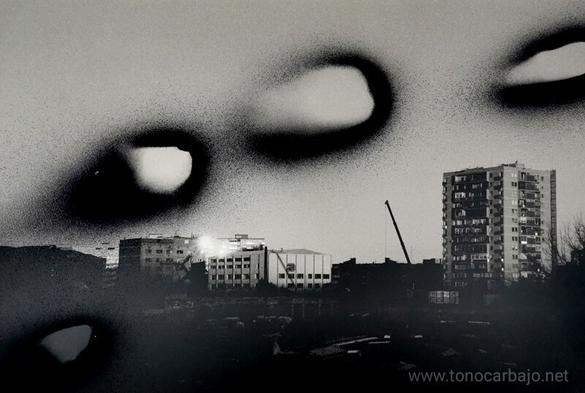 La imagen es una fotografía en blanco y negro que presenta una composición urbana con un elemento abstracto superpuesto. En la parte inferior, se observa un paisaje urbano con edificios residenciales, que se extienden a lo largo del horizonte. La fotografía fue tomada durante la puesta de sol. Una ventana refleja un destello de luz.
Lo más distintivo de la imagen son las formas abstractas y orgánicas que flotan sobre el paisaje. Estas formas, a spray de color negro intenso con bordes difuminados, crean un contraste marcado con el fondo más claro y estructurado de los edificios. Su disposición sugiere movimiento y dinamismo, y su naturaleza abstracta invita a la interpretación subjetiva.

En la esquina inferior derecha de la imagen, se encuentra una dirección web: www.tonocarbajo.net. Esta inclusión sugiere que la imagen es obra de un fotógrafo o artista visual que utiliza esta plataforma para mostrar su trabajo. La presencia de esta URL también proporciona un contexto adicional, indicando que la imagen forma parte de un portafolio o colección en línea.

En general, la imagen combina elementos de fotografía urbana con abstracción, creando una composición visualmente intrigante y evocadora. La técnica del blanco y negro, junto con la superposición de formas abstractas, confiere a la imagen un carácter distintivo y artístico.