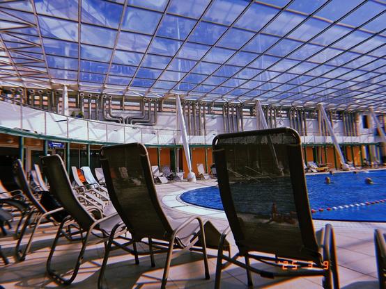 A row of reclining chairs next to a Swimmingpool. Glass roof with blue sky.