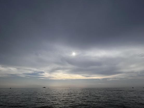The photo shows the sea under a cloudy sky. The sea and sky are nearly grey, towards the horizon the blue sky is visible. Through the clouds, in the middle of the photo the sun is visible as a bright dot.