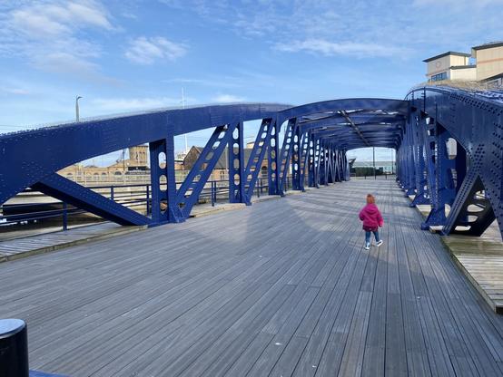 A toddler wearing a pink jacket and orange hat walking over the wooden surface of the Victoria Swing Bridge in Leith, Scotland. The metal structure of the bridge is painted a striking blue.