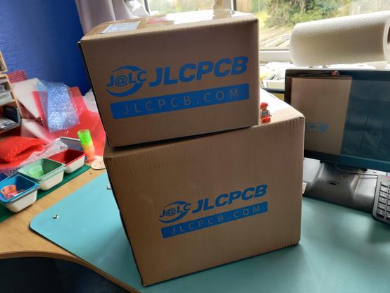 Two big brown cardboard boxes sigging on a workbench. The boxes are branded with the JLCPCB logs. A Lego Minifig sits on one of the boxes and looks tiny to indicate the scale.