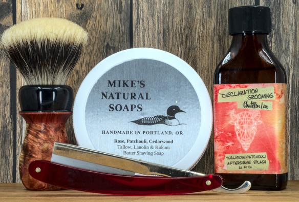 (Le Chat Mistral description)

This image features grooming items arranged on a wooden surface. On the left, there's a shaving brush with a wooden handle and white bristles. In the centre, there's a round tin labelled "Mike's Natural Soaps," depicting a duck and listing scents like rose and cedarwood. To the right, a brown glass bottle contains "Declaration Grooming" aftershave splash with yuzu, rose, and patchouli scents. In front, a red-handled straight razor lies horizontally. The background is rustic wood, giving a vintage, masculine feel.