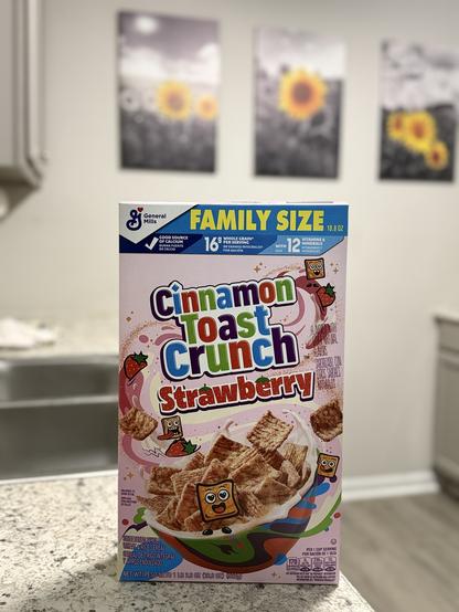 A box of Cinnamon Toast Crunch Strawberry cereal sits on a speckled gray countertop, in front of a sink and cabinets. Behind the cereal box on the wall are three framed pictures that are mostly black and white, but have sunflowers in them. The cereal box is pink and has the words "Cinnamon Toast Crunch Strawberry" in large, colorful letters on it. There are images of strawberries and Cinnamon Toast Crunch cereal pieces with faces on the box. The top of the box is blue and says "Family Size".