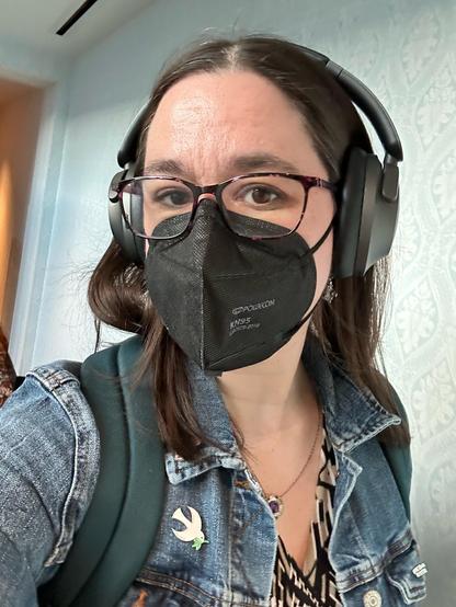 Selfie of me in a mask and big glasses and headphones. I’m wearing a blue jean jacket over a dress. The jean jacket has a dove pin.