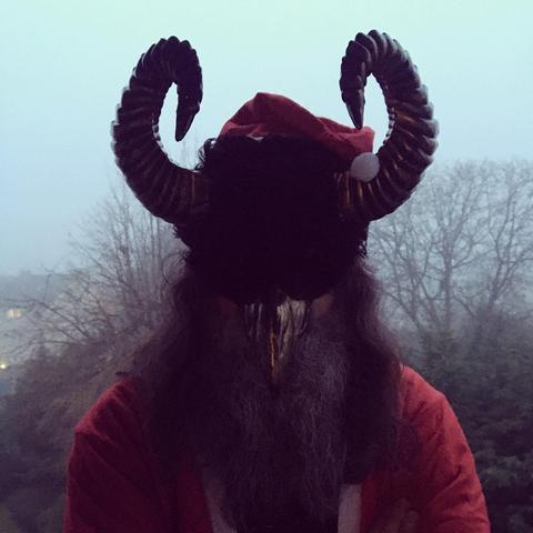 Ein düsteres Foto zeigt eine Person mit einer Krampus-Maske, die einen roten Weihnachtsmannmantel mit passender Mütze trägt. Die Maske ist schwarz und hat große, gerippte Hörner, die sich nach oben und leicht nach außen biegen; außerdem einen großen, goldenen Schnabel. Lange, dunkle Haare und ein grober Bart bedecken den Hals und die Brust der Person. Im Hintergrund ist eine neblige Landschaft mit Bäumen und Gebäuden zu sehen. Die Szene wirkt unheimlich und weihnachtlich zugleich.