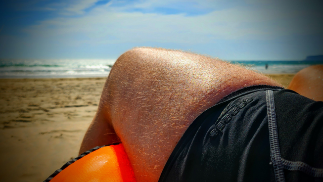 Jemand sitz am Strand in Badehose. Es ist sonnig, auf dem Strand ist viel Plarz und das Meer glänzt im Sonnenlicht.
