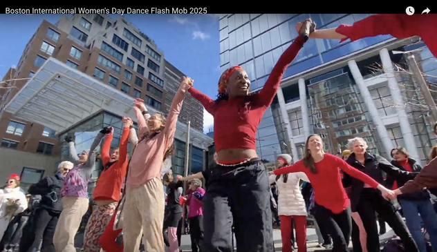 Still frame from the linked video of a large flash mob of women, all ages, races, ethnicities, genders, wearing varying shades of red and pink. They are dancing with great joy and energy outside on Boston's Boylston street. Arms raised, holding hands, and with huge smiles as they greet the thousands of of International Women's Day marchers coming down street.