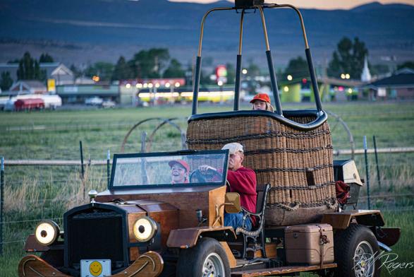 An old-fashioned looking truck hauls the wicker basket for a hot air balloon to the Panguitch, Utah Balloon Rally. One child sits next to the driver, while a young girl rides inside the basket attached to the truck bed.