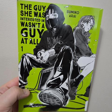 Photograph of a white person's hand holding up The Guy She Was Interested In Wasn't a Guy At All, volume 1 against a light colored wall. The cover has the title in all capital, black lettering along the left side of the page with a neon green background. The cover has a person centralized sitting on an amp wearing baggy pants, a hoodie, and a face mask to cover their mouth. Sitting on the other side of the amp is a blushing, feminine character in a school uniform with a skirt.