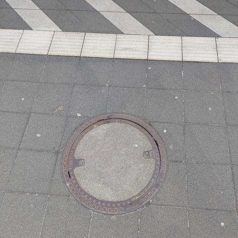 A round manhole cover made from metal and concrete in the middle of grey, square stone tiles. Next to it there is white colored tactile paving.
