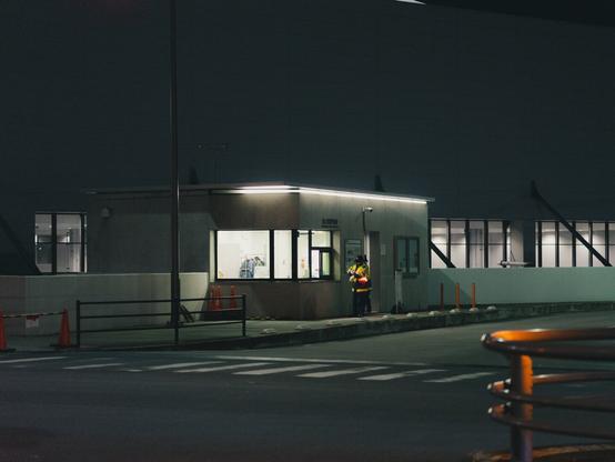 The image shows a small building or security booth at night, illuminated by lights. There appears to be a person wearing a reflective vest standing near the entrance. Traffic cones and barriers are present nearby, and the building is adjacent to a road with pedestrian crossings visible in the foreground.