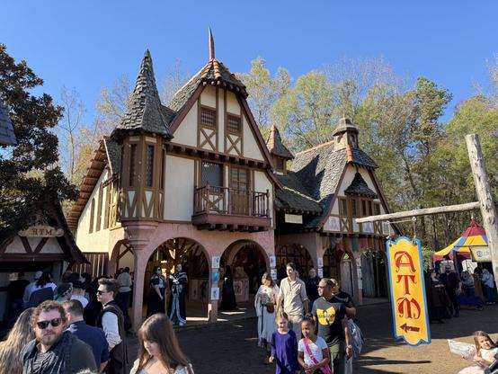 A medieval-style building at a Renaissance Festival with costumed attendees walking by, an ATM sign, and a vibrant market atmosphere.