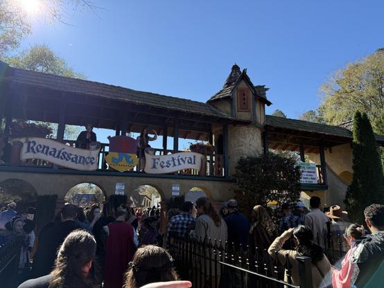 A crowd gathers at the entrance of a Renaissance Festival, with costumed performers standing on a medieval-style bridge under a clear blue sky.