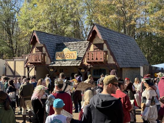 A lively crowd in medieval attire gathers outside a rustic bakery at a Renaissance Festival, with people chatting and ordering food.