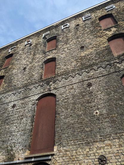 Façade latérale régulièrement percée d'ouvertures condamnées de tôles. En haut, les lettres blanches de la fin du nom de l'industriel : FRÈRES.