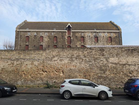 Derrière un haut mur de briques, la façade latérale du bâtiment. En lettres blanches : SAINT FRÈRES.