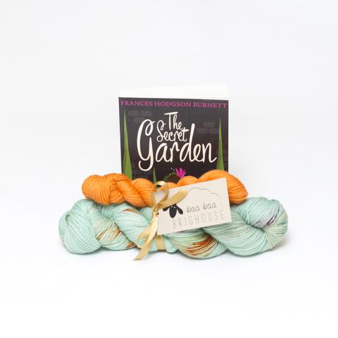 Picture shows a sock set of yarn consisting of a main skein which is pale green with speckles of deep orange, purple and green and a mini skein which is orange. The two are tied together with gold ribbon. Behind the yarn, stood on its end is a copy of the book, The Secret Garden.