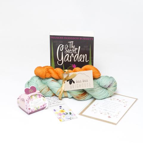 Picture shows a sock set of yarn consisting of a main skein which is pale green with speckles of deep orange, purple and green and a mini skein which is orange. The two are tied together with gold ribbon. Behind the yarn, stood on its end is a copy of the book, The Secret Garden. In front of the yarn is a gift box of soaps, a bookmark and a card.