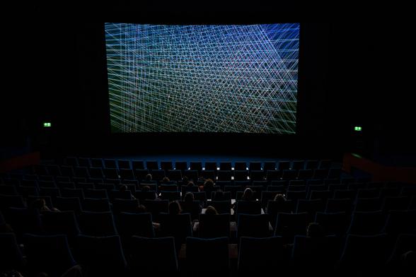 A dark cinema with some people visible. The screen shows an abstract, colourful pattern of many intersecting lines.